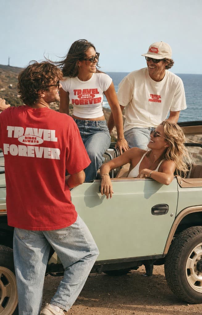 Four people posing with a mint-green vintage open-top off-road vehicle on a sunny coastal overlook, wearing Travel Forever shirts.