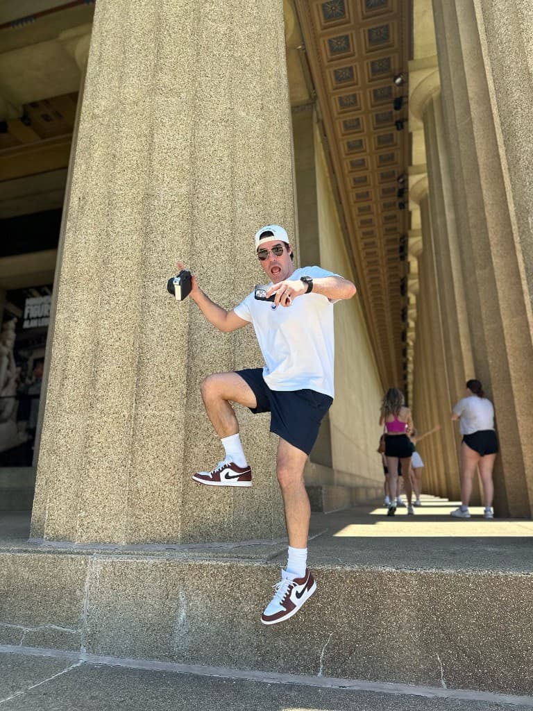 A man jumping between stone columns with a camera in one hand and a phone in the other.