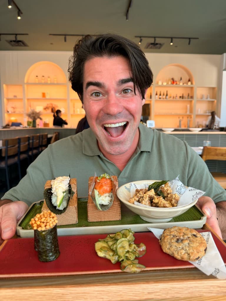 An enthusiastic man holding a wooden tray of handrolls, fried karaage, and a chocolate chip cookie at Cuddlefish in Atlanta.